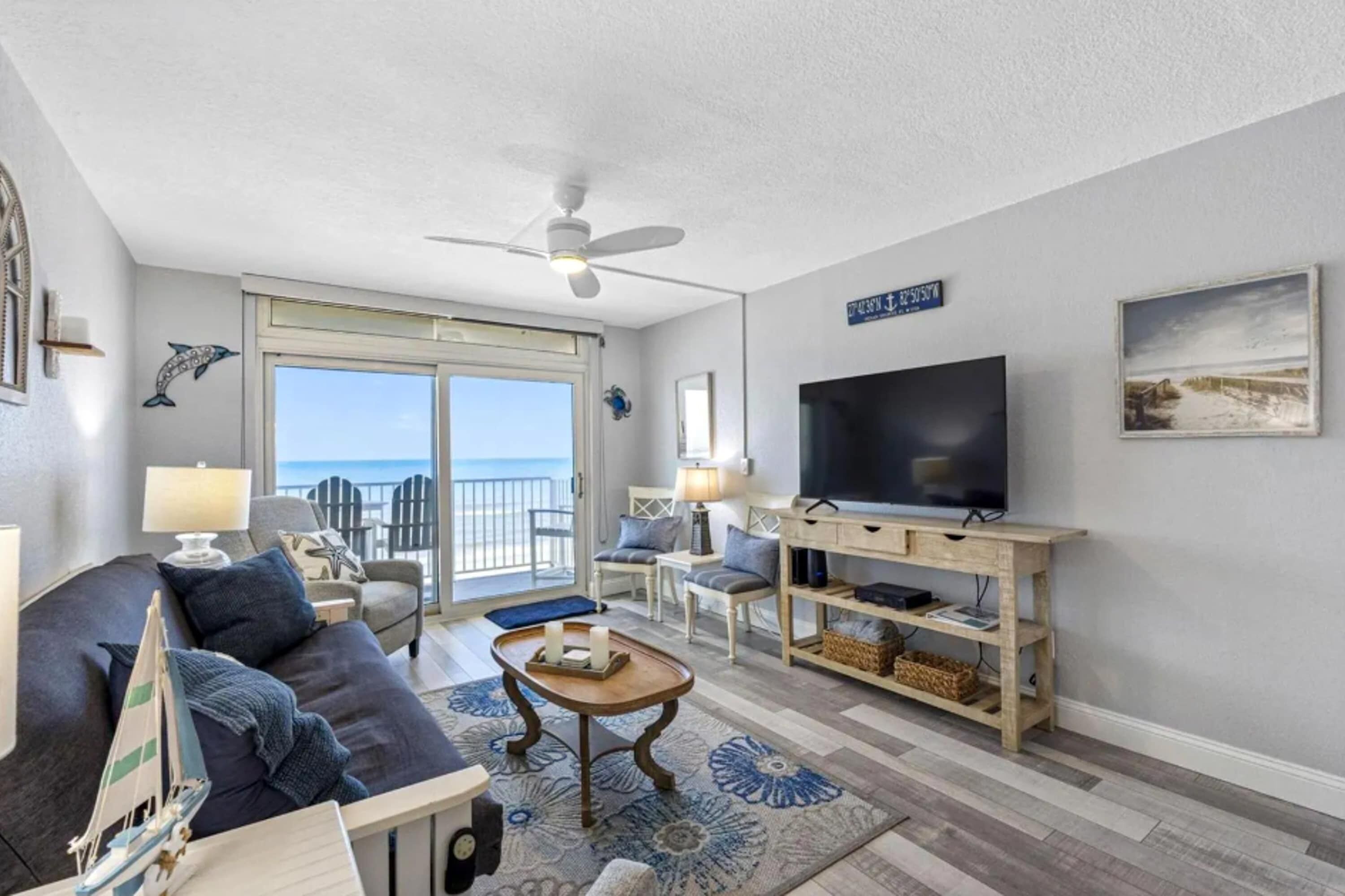 A living room with a sofa, coffee table, TV, and a view of the ocean through a sliding glass door.