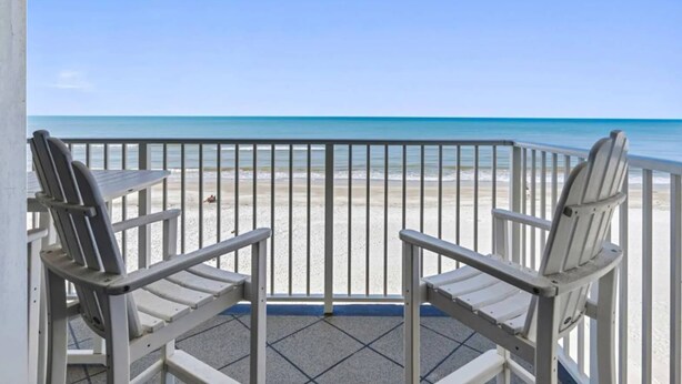 Ein Balkon mit zwei weißen Stühlen, der auf einen Strand blickt.
