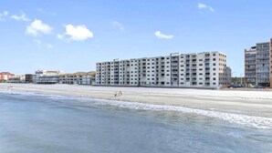 Ein Hotel direkt am Strand mit mehreren Etagen, ein sandiger Strand im Vordergrund und ein klarer Himmel.