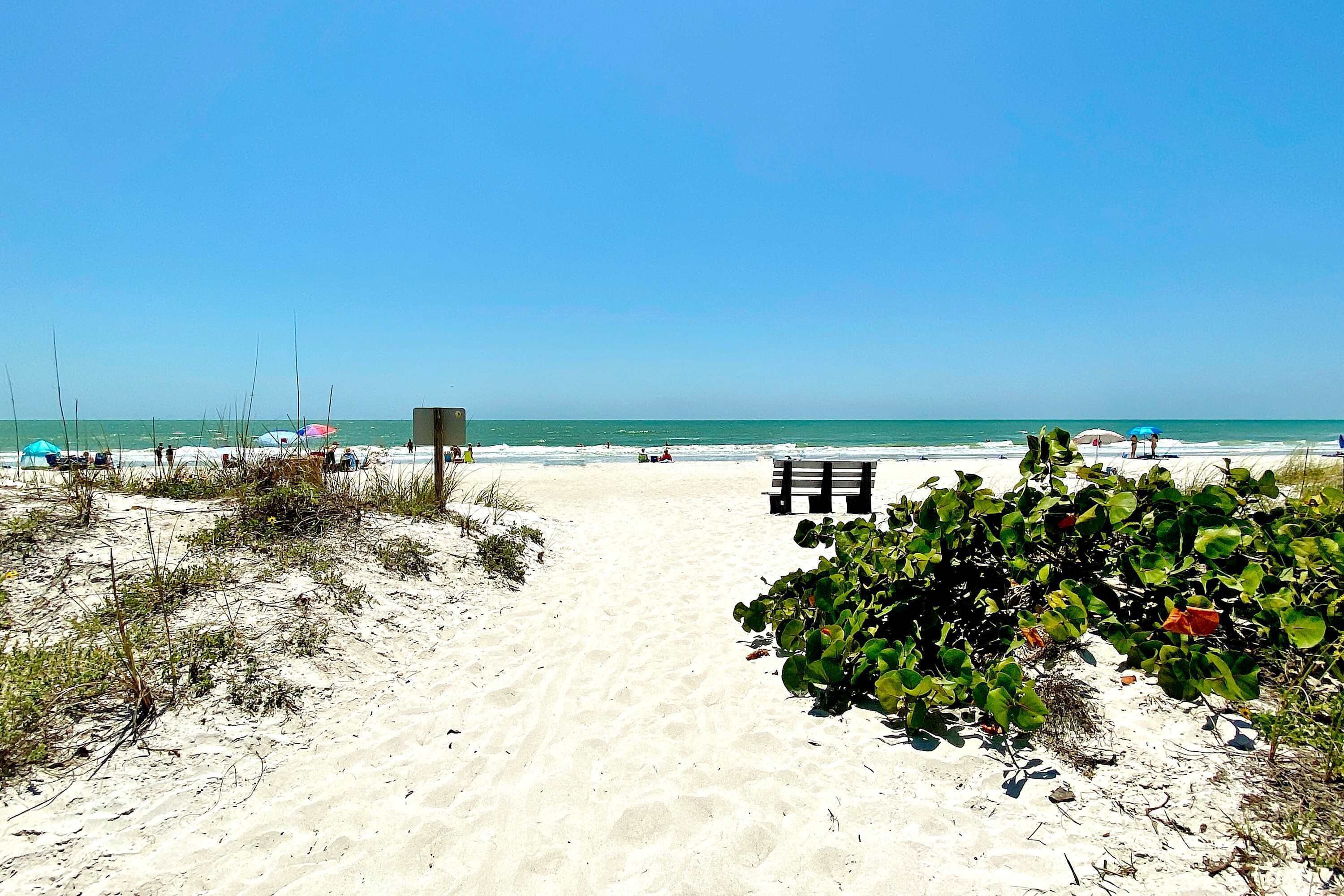 A sandy beach path leading to the sea with sparse vegetation and a clear sky.