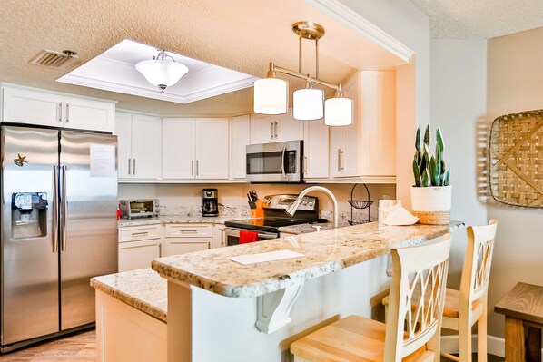 A modern kitchen with white cabinets, stainless steel appliances, a granite countertop, and pendant lighting. - 4th floor beachfront corner condo with private balcony views BBQ AC (Indian Shores)