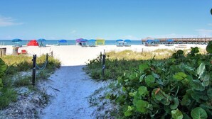 A beach path leading to a sandy beach with umbrellas and a wooden pier in the background.