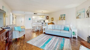A spacious living room with a light blue sofa, patterned armchair, and a wooden coffee table. A kitchen area is visible in the background with white cabinets and a dining table. - Charming condo with gorgeous Gulf views and beachfront heated pool and spa (Indian Shores)