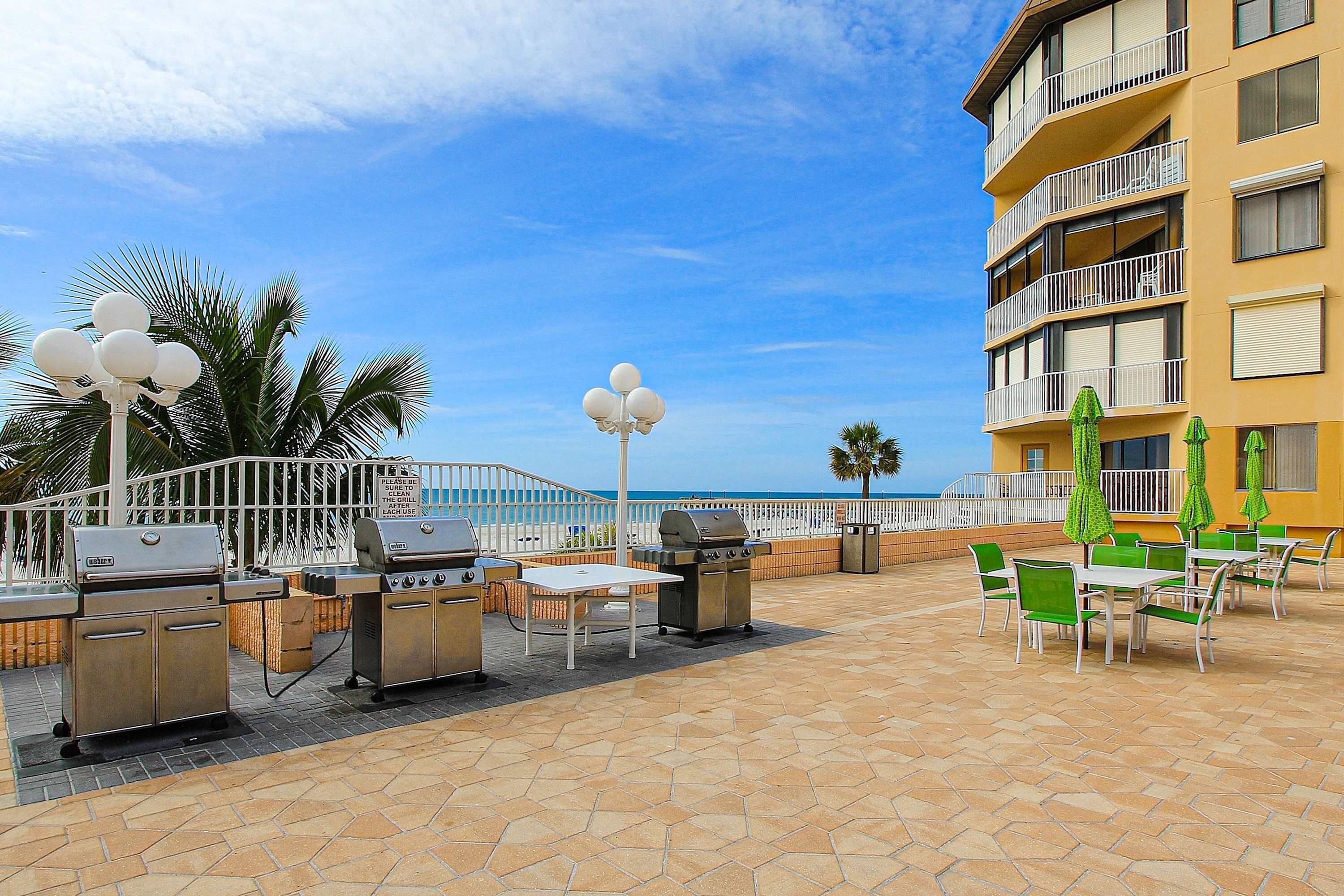 Un patio exterior con parrillas, mesas y sillas, con vista a una playa y un edificio amarillo con balcones.