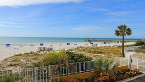 A beach with blue lounge chairs, a palm tree, and a pier in the background. - Charming condo with gorgeous Gulf views and beachfront heated pool and spa (Indian Shores)