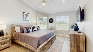 Un dormitorio con una cama de madera, un ventilador de techo, un televisor montado y vistas al exterior a través de amplias ventanas.