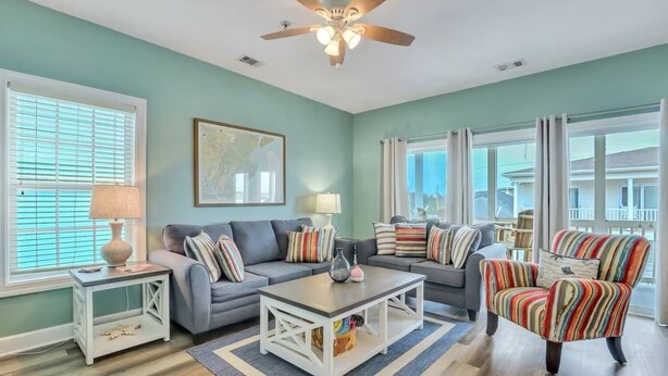 A living room with a grey sofa, striped armchair, and a coffee table. There are two lamps on side tables, a ceiling fan, and a large window with blinds. - Lovely Tybee Condo Close to Beach (Tybee Island)