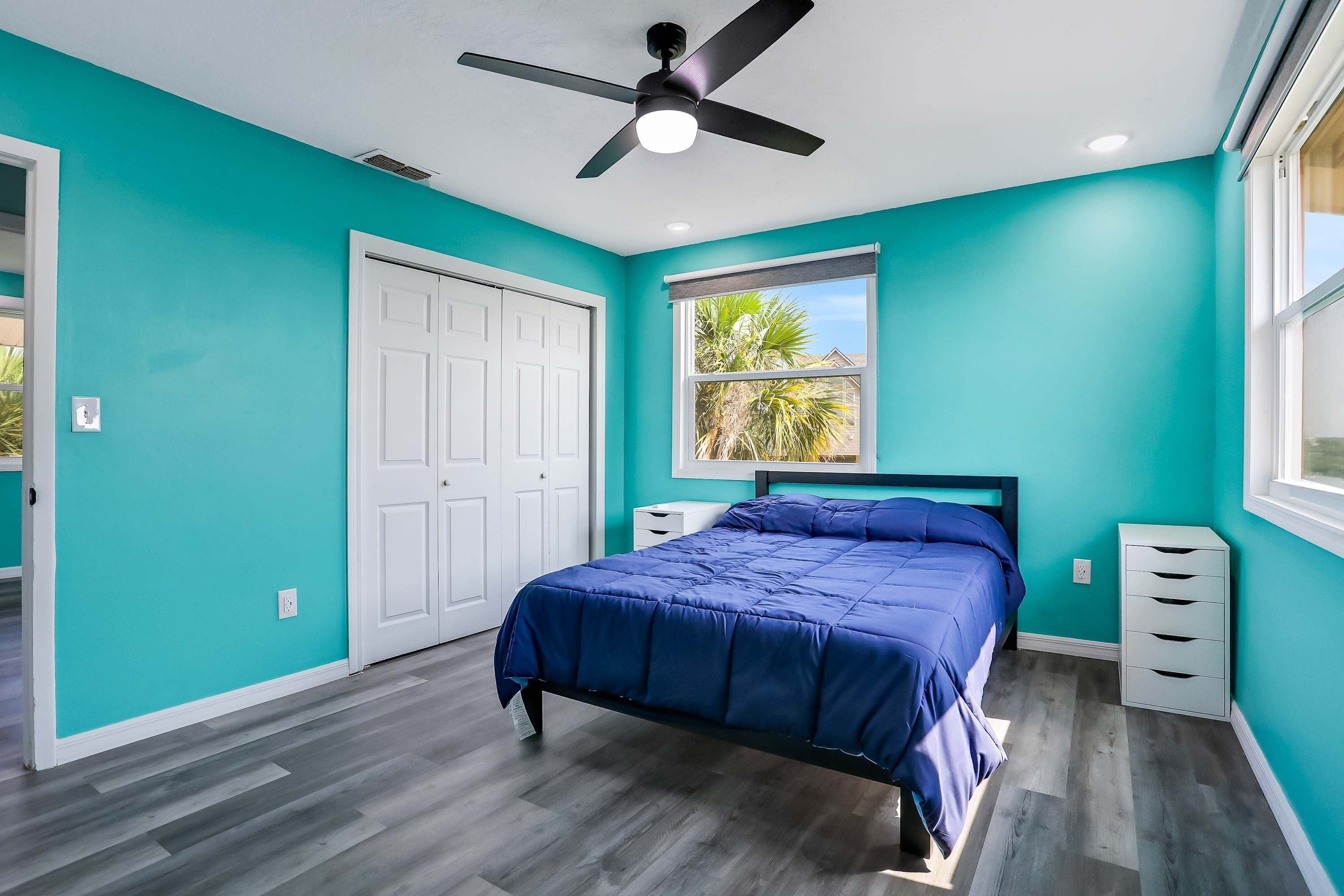 A bedroom with teal walls, a bed with a blue bedspread, a ceiling fan, a window with a view of palm trees, and a white door.