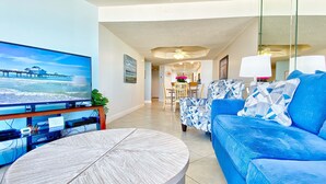 Ein Wohnzimmer mit einem großen Flachbildfernseher, einem blauen Sofa, einem runden Couchtisch und einem Essbereich im Hintergrund.