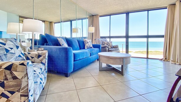 Ein modernes Hotelzimmer mit großem Fenster und Meerblick, einem blauen Sofa, einem gemusterten Sessel und einem kleinen runden Tisch.