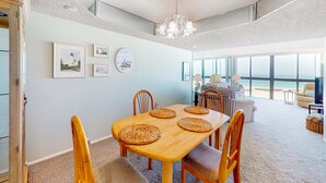 A dining area with a wooden table, four chairs, and woven placemats. There are framed pictures on the wall, a chandelier, and a view of the beach through large windows.