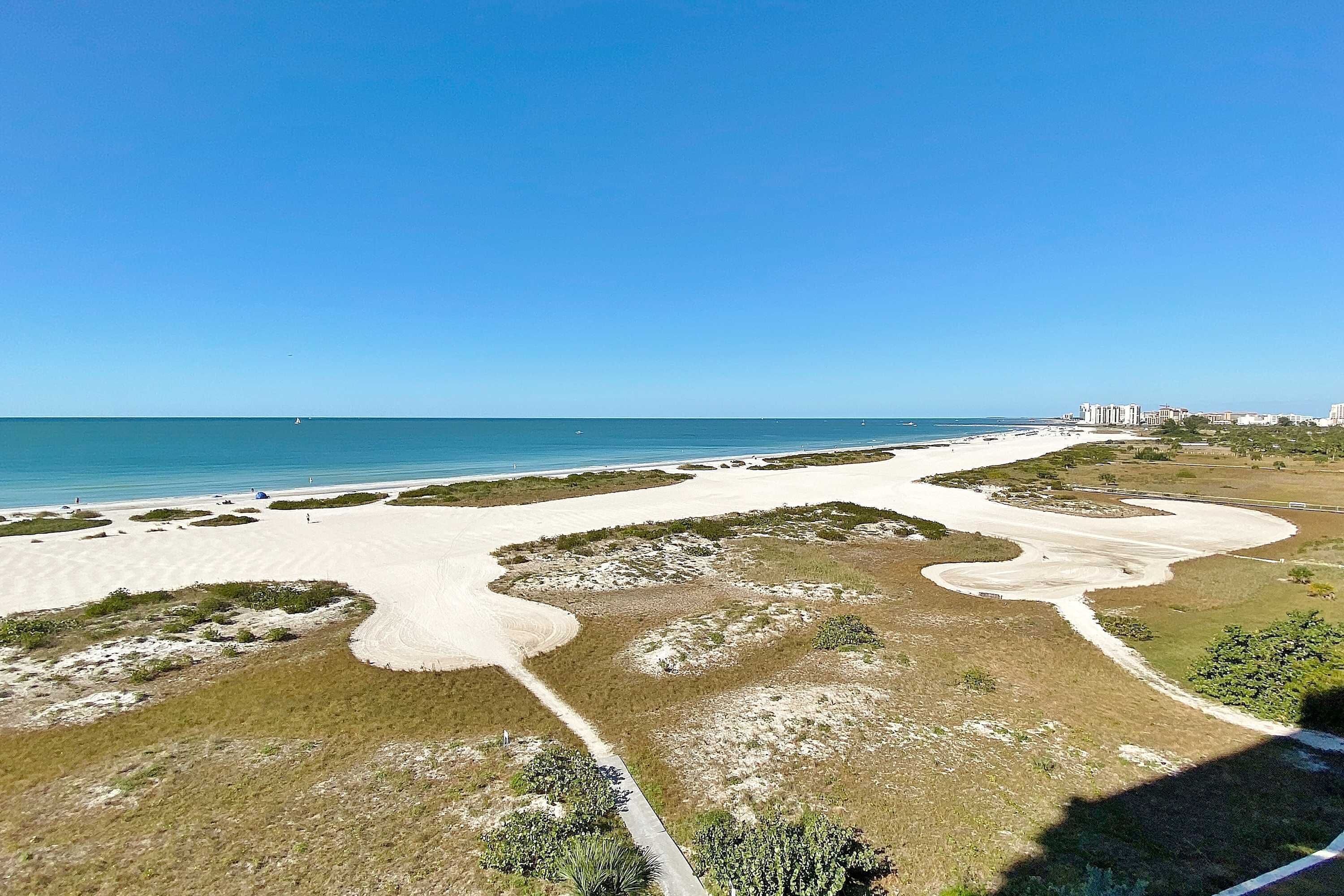 Un paisaje costero con una playa arenosa, un sendero serpenteante y un horizonte urbano distante.