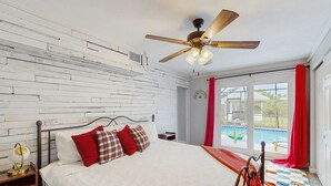 Une chambre à coucher avec un lit, une tête de lit en bois, un ventilateur de plafond, une table de chevet et une vue sur une piscine, visible à travers une grande fenêtre.