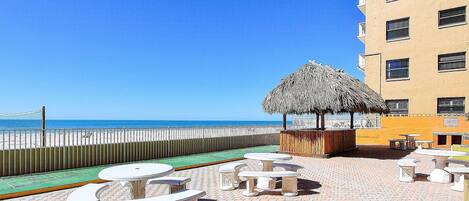 Un patio frente a la playa con mesas y sillas, una sombrilla de paja y vista al océano.