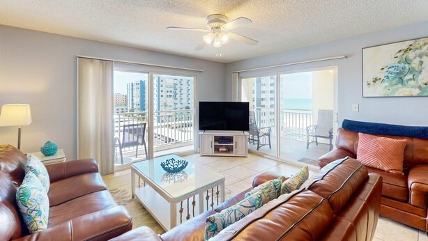 A spacious living room with a ceiling fan, a flat-screen TV, and a balcony with a view of the city.