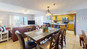 A modern kitchen with wooden cabinets, a microwave, and a refrigerator. There is a dining area with a table and chairs, and a living room with a sofa and a TV.