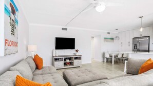 Een moderne woonkamer met grijze banken en oranje kussens, een flatscreen televisie en een keuken op de achtergrond.