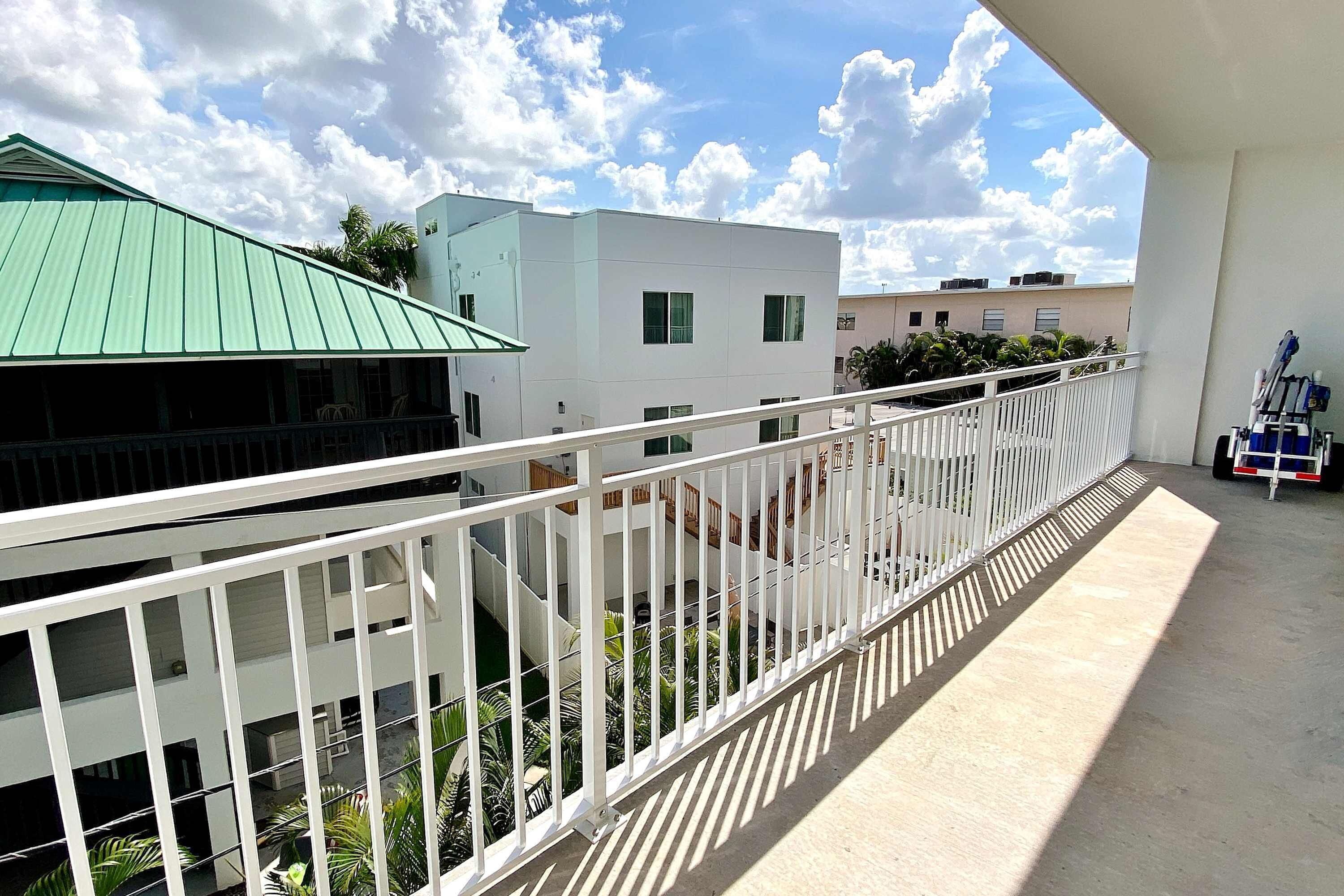 A balcony with a white railing overlooking a courtyard with greenery and buildings in the background.