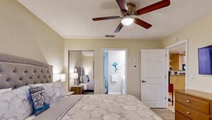 Een slaapkamer met een gestoffeerd hoofdeinde, een groot bed met beddengoed met een patroon, een plafondventilator en een houten dressoir.