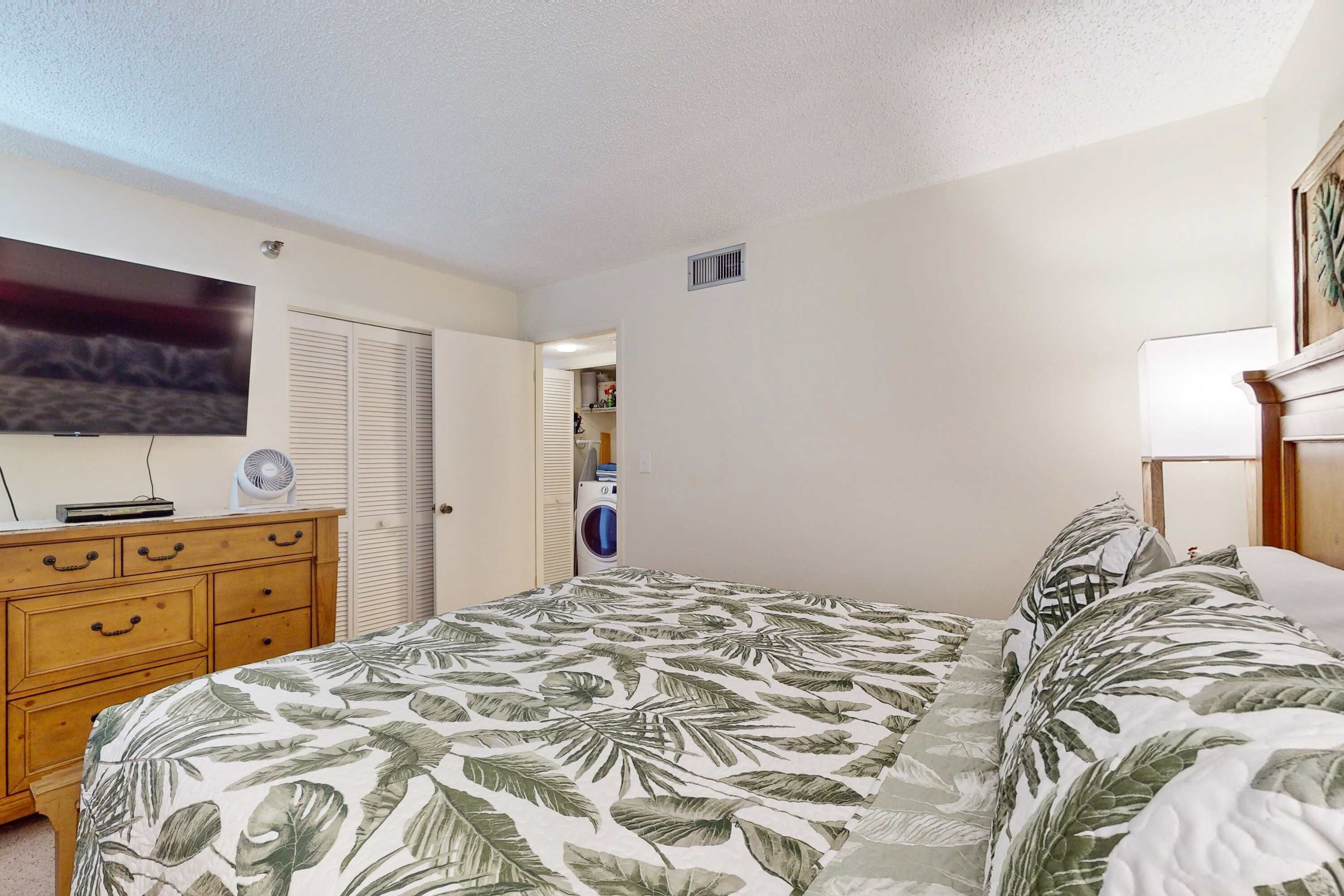 A bedroom with a bed, wooden dresser, TV, and a washing machine in the background.