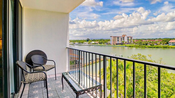 A balcony with two chairs and a table, overlooking a river with buildings in the background. - Waterfront condo with pool and spa directly across from wide beach (Indian Shores)