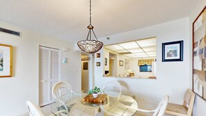 A dining area with a glass table surrounded by white chairs, a hanging light fixture, and framed pictures on the walls. - Waterfront condo with pool and spa directly across from wide beach (Indian Shores)