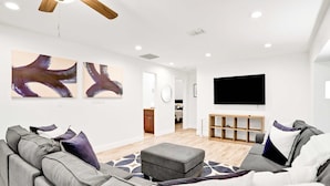 Un salon moderne avec un ensemble de canapés gris, un pouf gris, des œuvres d’art abstraites accrochées au mur, un téléviseur à écran plat et un meuble de rangement en bois pour les appareils multimédias.