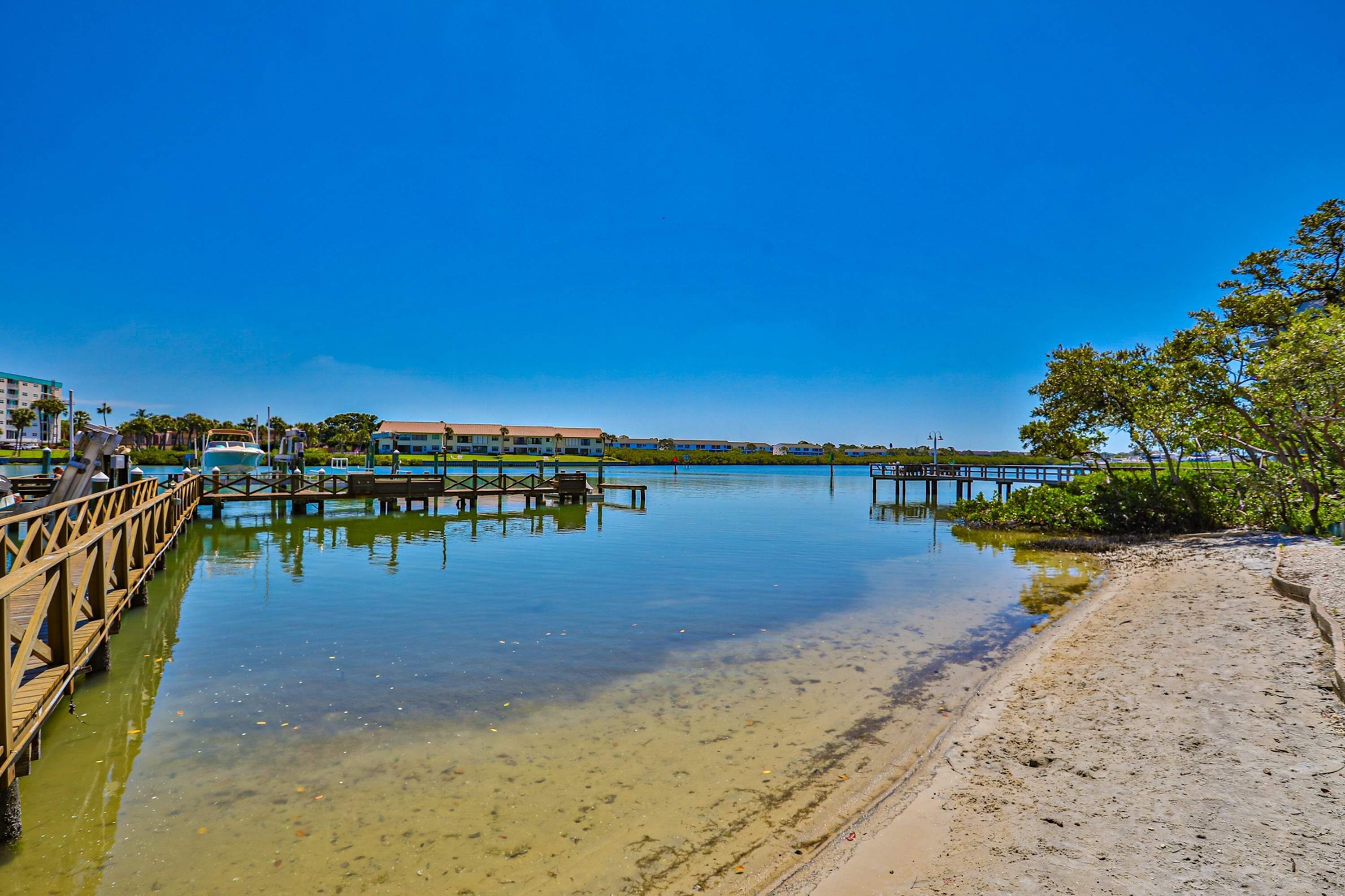 A waterfront view with a wooden pier extending into calm water, a sandy beach, and a clear blue sky.