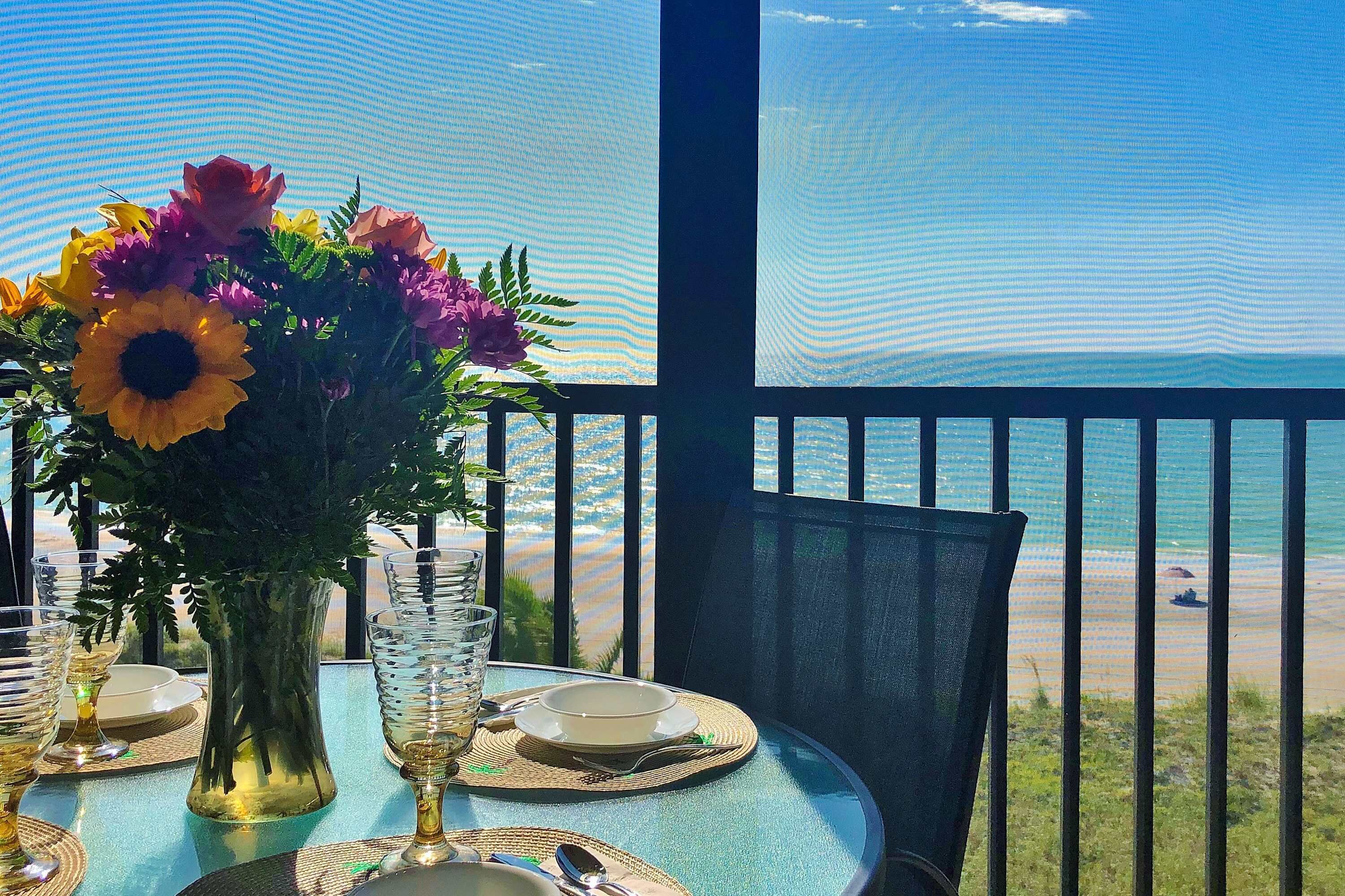 Mesa posta com um vaso de flores, pratos e talheres, com vista para uma praia.