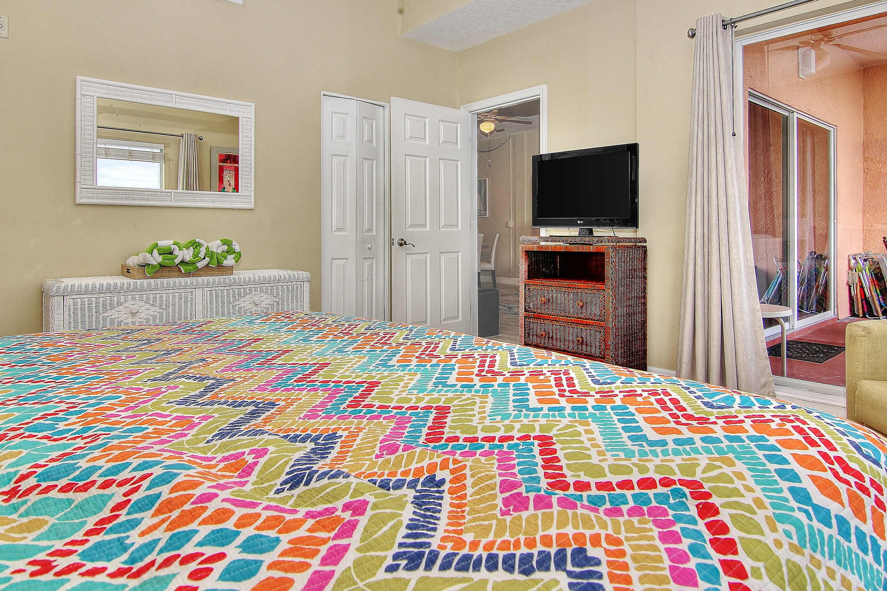 A bedroom with a colorful quilt, a television on a stand, and a door leading to another room.