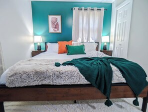 A bed with a fluffy white cover, a green blanket, and two pillows in a room with teal walls, white curtains, and a framed picture on the wall.