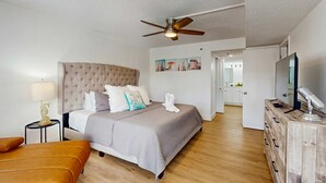 Une chambre d’hôtel avec un lit doté d’une tête de lit capitonnée, un plancher en bois, un ventilateur de plafond, une télévision et une commode en bois.