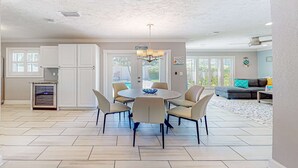 A spacious dining area with a round table and six chairs, a kitchen with white cabinets, and a large window providing natural light. - Dog friendly home near beaches with private pool enclosed backyard grill WD (St. Pete Beach)
