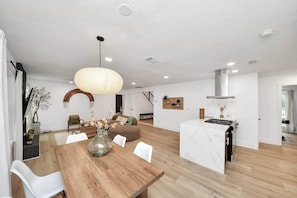 A spacious living area with a wooden dining table, a large hanging light fixture, and a kitchen with a marble countertop.