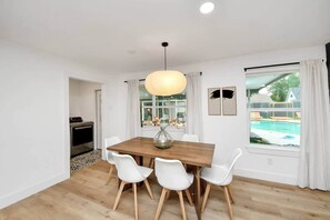 A dining area with a wooden table, white chairs, a hanging light fixture, a window with curtains, and a view of a swimming pool.
