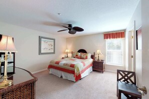 A bedroom with a bed, a ceiling fan, a nightstand, a chair, a lamp, a picture frame, and a window with blinds.