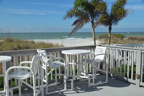 A deck with white outdoor furniture overlooking a beach with white sand and ocean waves.