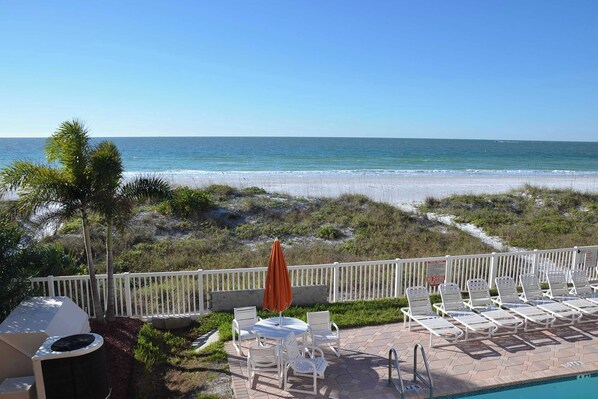 A poolside area with white lounge chairs and an umbrella, overlooking a beach with white sand and a clear blue sky.