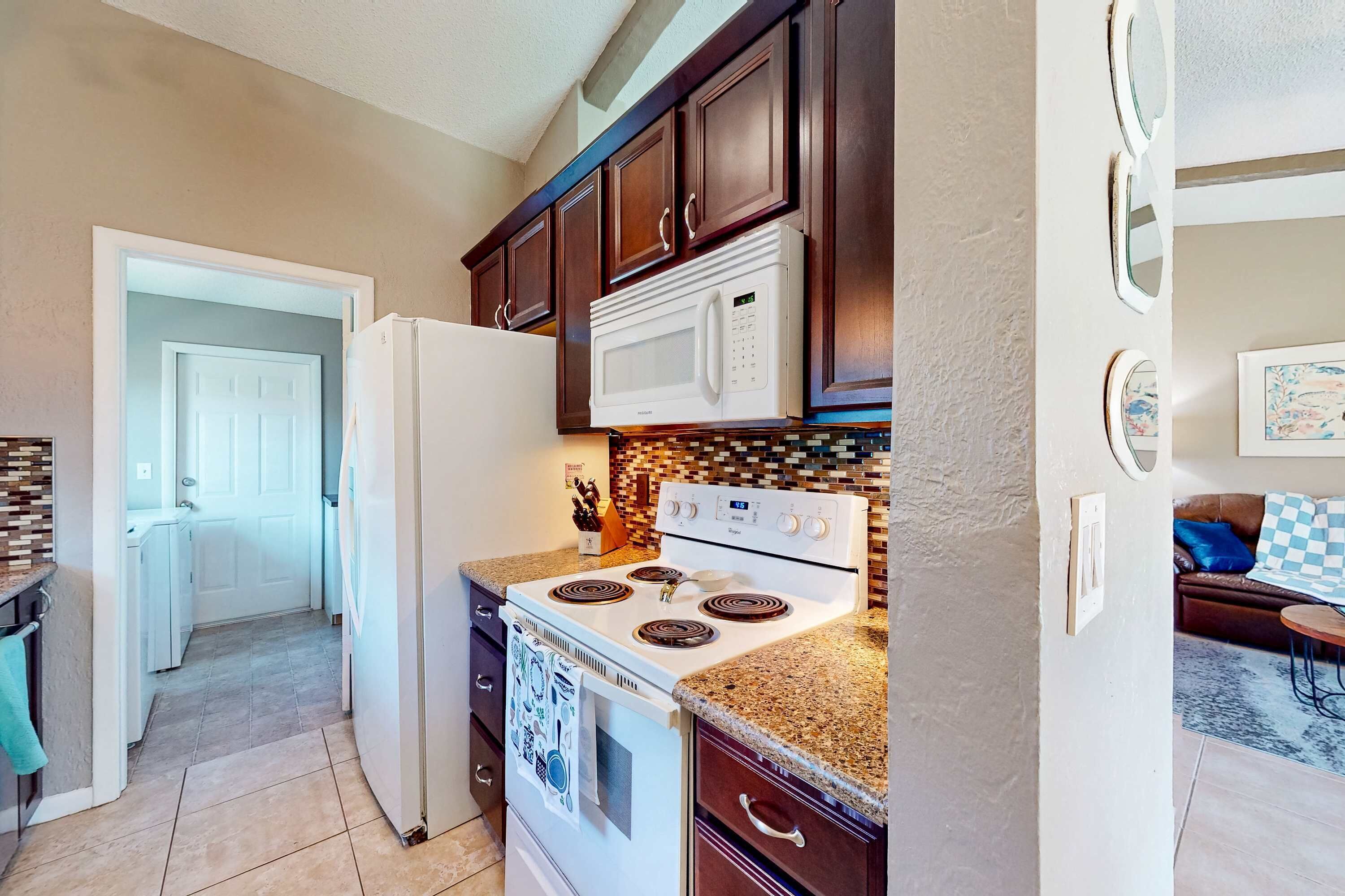 Una cocina con armarios de madera oscura, un refrigerador blanco, una estufa blanca y un microondas. Cuenta con un backsplash de azulejos y una puerta que da a otra habitación.