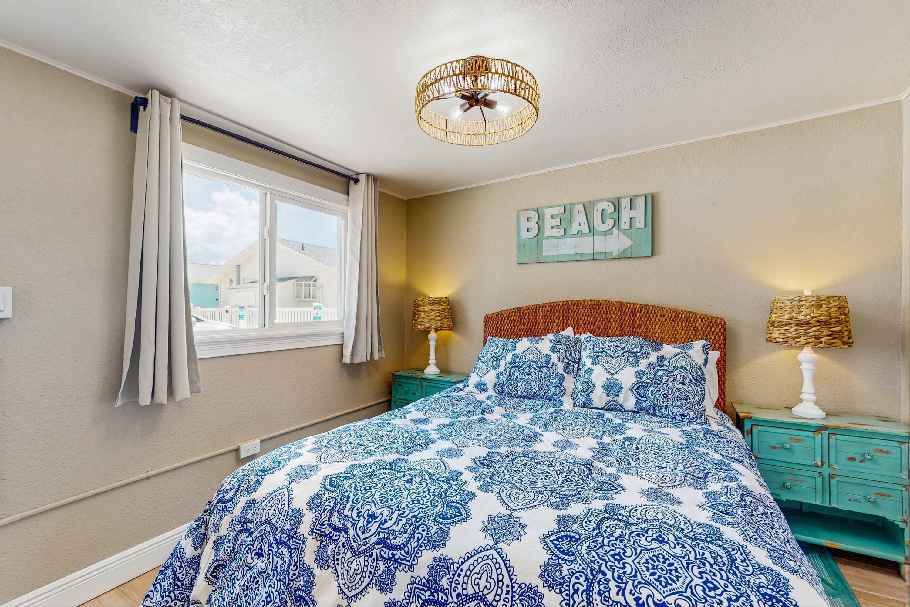 A bedroom with a large bed, a window with curtains, a wall-mounted 'BEACH' sign, and two bedside lamps.