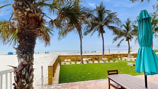 Un espace patio avec une table et des chaises en bois, une pelouse verdoyante et une vue sur la plage et les palmiers.
