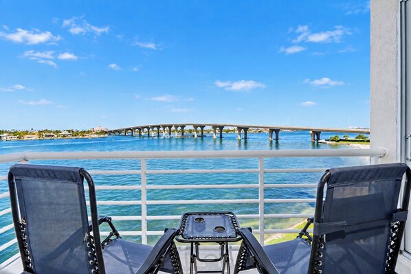 A balcony with two chairs and a small table, overlooking a body of water with a bridge in the distance.