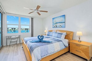 A hotel room with a bed, a desk, a chair, a nightstand, a lamp, a ceiling fan, a window with a view of a bridge, and a painting on the wall.