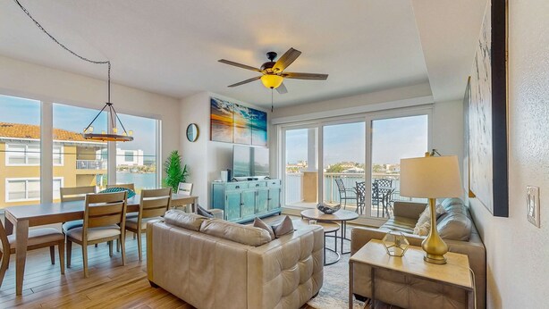 A modern living room with a sofa, dining table, and chairs, a ceiling fan, and large windows offering a view of the city. - Waterfront Condo w WiFi Washer Dryer and Central AC Snowbird Friendly (Clearwater Beach)