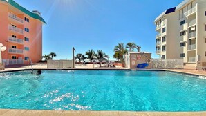 A swimming pool with clear blue water, surrounded by buildings with balconies and palm trees. - Dreamy condo with panoramic Gulf vIews and beachfront heated pool and spa (Indian Shores)