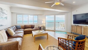 A spacious living room with a large sofa, a coffee table, and a television. There is a balcony with a view of the ocean. - Dreamy condo with panoramic Gulf vIews and beachfront heated pool and spa (Indian Shores)