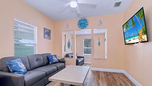 Un salon avec un canapé gris, une table basse en bois, un ventilateur de plafond, une horloge murale et un téléviseur diffusant une scène de plage.