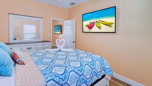 Une chambre avec un lit, un ventilateur de plafond, un tableau représentant des kayaks sur une plage, et une porte devant laquelle est posée une serviette en forme de cœur.