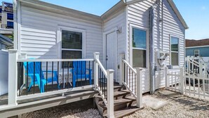 Une petite maison de plain-pied avec un porche, un escalier menant à la porte d’entrée et un petit espace clos agrémenté de chaises bleues.