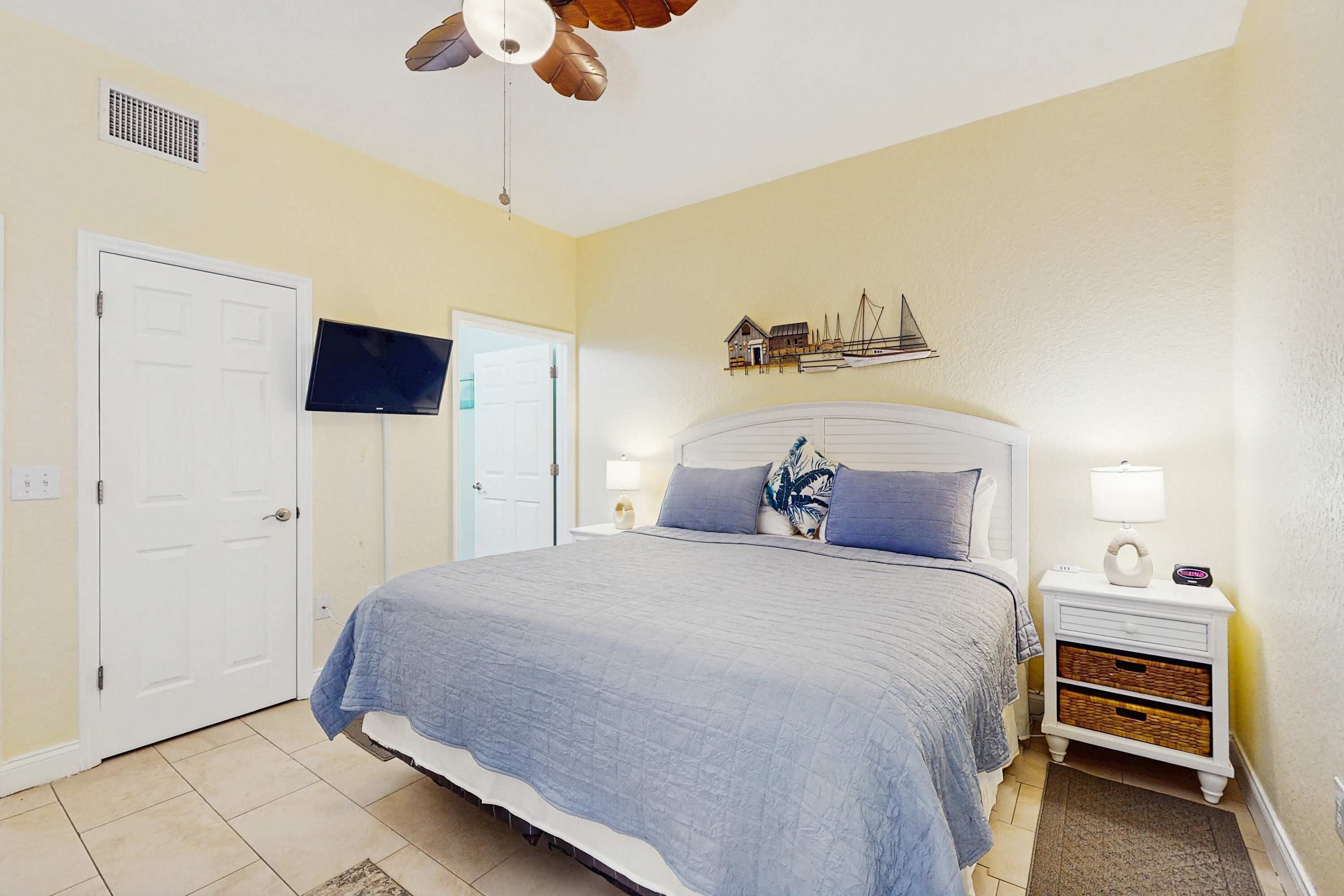 Un dormitorio con cama, mesita de noche, televisor montado en la pared, ventilador de techo y un adorno en la pared.